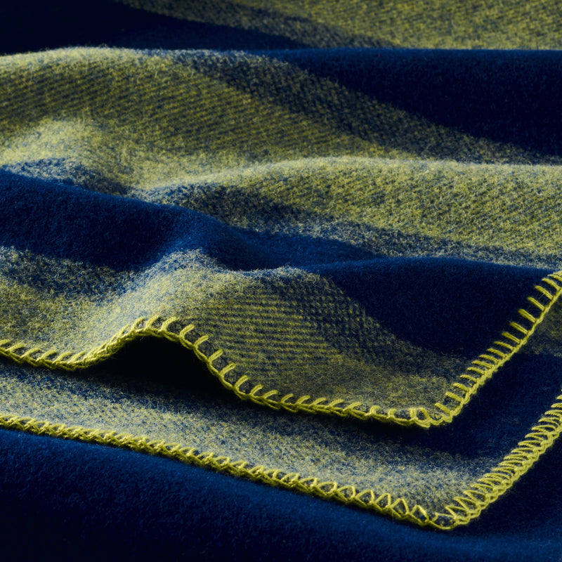 A closeup of a wool blanket showing the wool surface texture, and whipstitched edge. The pattern is a green and navy blue stripe and the edge stitching is green.